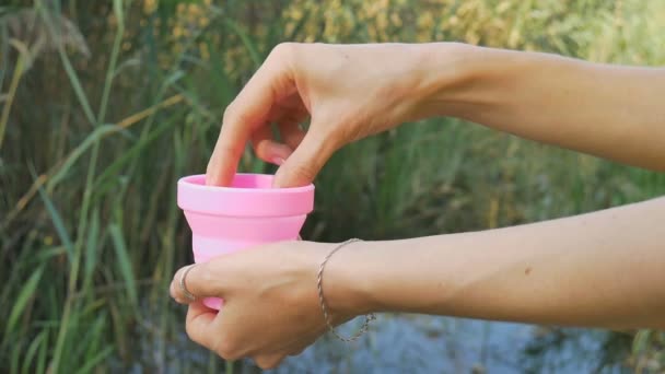 Les mains féminines montrent comment utiliser une coupe menstruelle en silicone rose debout sur fond naturel vert avec lac. Concept de santé des femmes, solutions zéro déchet 