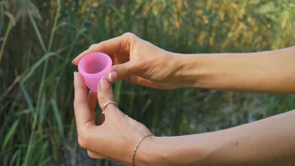 Woman holding a pink menstrual cup standing against green natural background with lake. Female hygiene concept — Stock Video