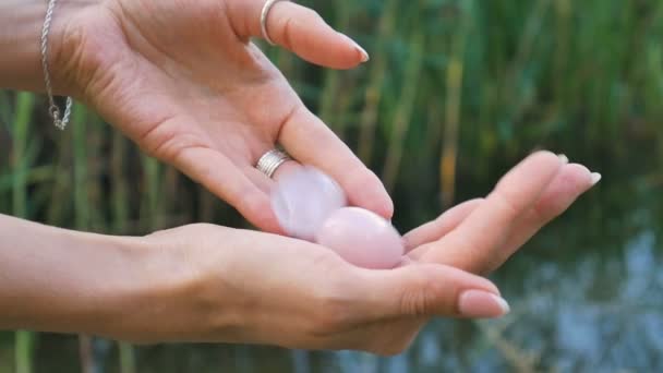 Női kéz kezében egy rózsa kvarc és ametiszt kristály yoni tojás a folyó hátterében. A nők egészsége, egység a természet fogalmaival — Stock videók