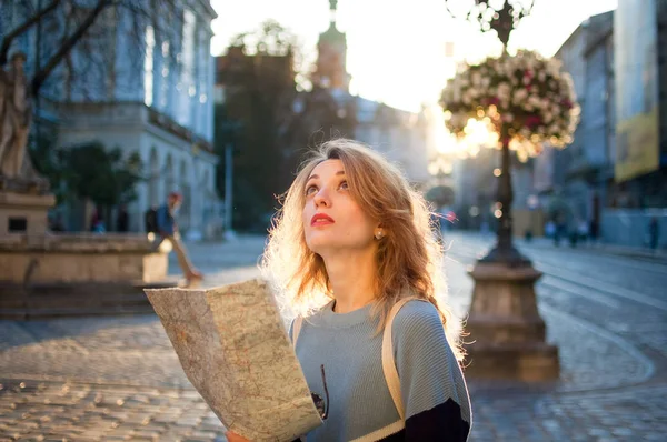 Счастливая взволнованная взрослая девушка смотрит на бумажную карту и ищет направление рано утром в древнем европейском городе на пустой площади — стоковое фото