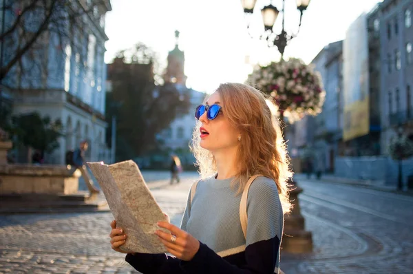 Улыбающаяся юная туристка в солнечных очках и бумажной карте в руках ищет направление ранним утром на пустой древней площади — стоковое фото