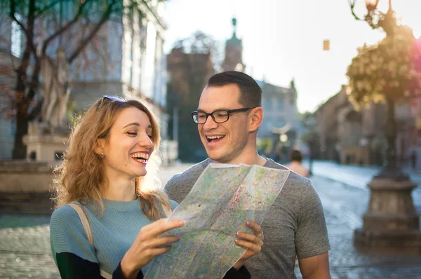 Young happy laughing couple on vacation in European city early in the morning, girl is holding a paper map in hands, man is wearing eyeglasses