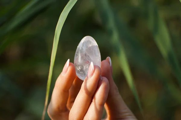 Mano femminile con manicure francese che tiene trasparente viola uovo ametista yoni per vumfit, imbuilding o meditazione. Uovo di quarzo cristallo in mano su sfondo verde all'aperto — Foto Stock