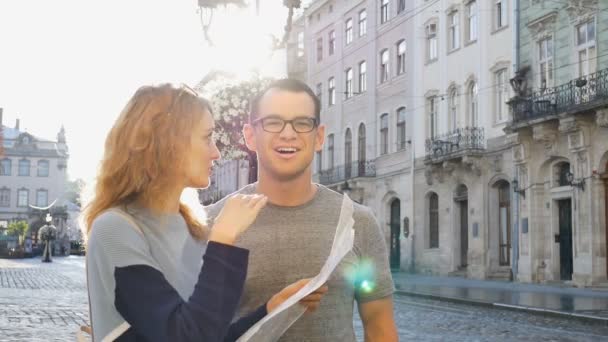 Feliz pareja de jóvenes turistas sosteniendo un mapa en papel de la antigua ciudad europea temprano en la mañana en la plaza vacía y señalando algún lugar turístico — Vídeos de Stock