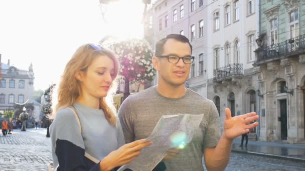 Lyckliga unga turister par som håller en papperskarta över gamla europeiska staden tidigt på morgonen på tomma torget och pekar på några sightseeing plats — Stockvideo