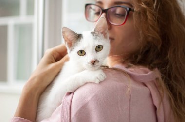 Pembe giysiler ve gözlük lü çekici kız kollarında kedi ile evde pencerenin yanında duran. Evcil hayvan sahibi kavramı