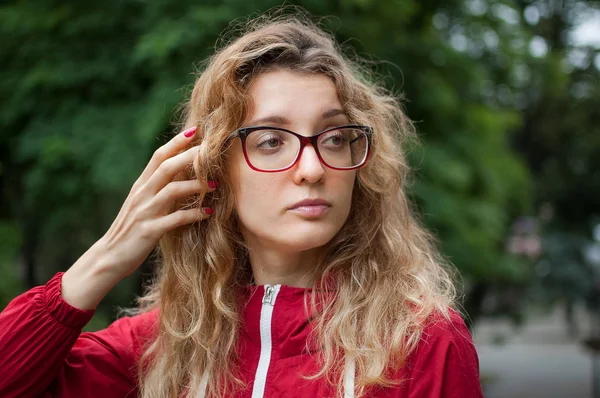 Junge schöne Frau mit blonden lockigen Haaren in dunkelroter Hipsterjacke und Brille posiert morgens im Stadtpark — Stockfoto