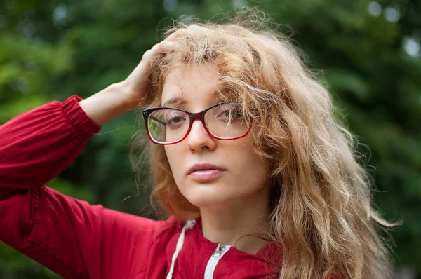 Junge schöne Frau mit blonden lockigen Haaren in dunkelroter Hipsterjacke und Brille posiert morgens im Stadtpark — Stockfoto