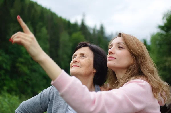 Feliz madre mayor y la hija adulta están viajando y posando juntos sobre el paisaje de bosque y montañas, niña está apuntando a algo. Concepto de viajar en familia —  Fotos de Stock