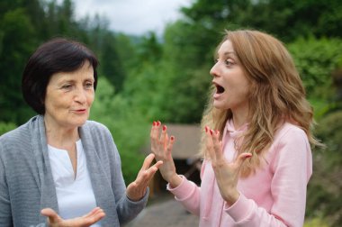 Outdoors portrait of senior mother and adult daughter are communicating outside over landscape of forest and mountains. Two generations, problems between children and parents concept clipart