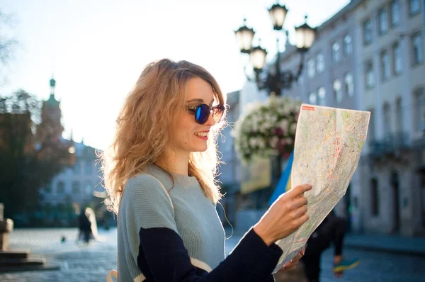 Menina adulta em óculos de sol azuis está olhando para o mapa de papel e procurando direção no início da manhã na antiga cidade europeia na praça vazia — Fotografia de Stock