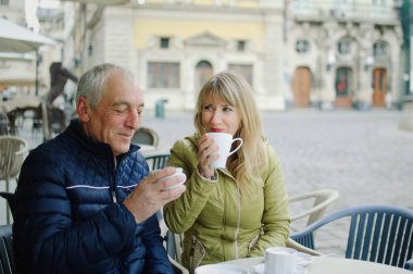 Portrait of happy romantic couple with age difference drinking coffee in cafe with terrace outdoors in the ancient city in the morning during early spring or autumn. clipart