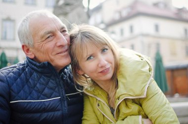 Yakışıklı yaşlı adam, genç sarışın karısını kucaklıyor. Baharın başında ya da sonbaharda antik şehirde birlikte vakit geçiriyorlar..