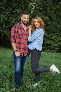 Açık havada, kırmızı gömlekli sakallı hippi erkek arkadaşın ve yeşil ağaçların arkasındaki parkta mavi günlük kot bluz giyen sarışın kız arkadaşın tam boy portresi.