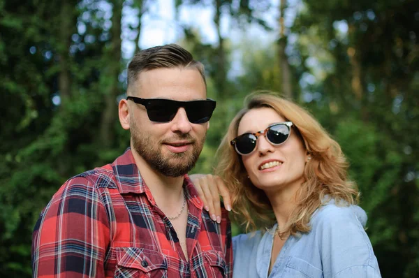 Lovely couple of bearded hipster boyfriend in red shirt and blond girlfriend in blue casual jeans blouse looking at the camera wearing dark sunglasses