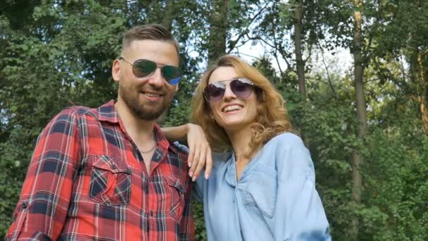 Lovely couple of bearded hipster boyfriend in red shirt and blond girlfriend in blue casual jeans blouse in dark sunglasses looking at the camera and smiling on green trees background — Stock Video