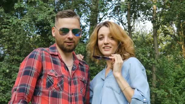 Charmant couple de hipster boyfriend barbu en chemise rouge et petite amie blonde en jean décontracté bleu regardant la caméra et enlevant leurs lunettes de soleil sombres sur fond d'arbres verts — Video