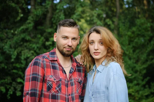 Lovely couple of bearded hipster boyfriend in red shirt and blond girlfriend wearing blue casual jeans blouse looking at the camera on green trees background