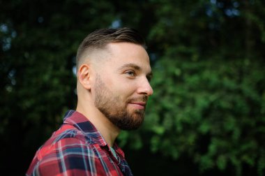 Kırmızı gömlekli vahşi sakallı genç adam dışarıda yeşil ağaçların arkasında duruyor. Hipster tarzı konsept.