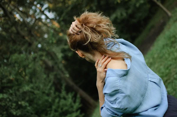 Вид сзади на взрослую блондинку в синих джинсовых блузках, позирующую в парке на зеленом фоне деревьев. Концепция стиля хипстера . — стоковое фото