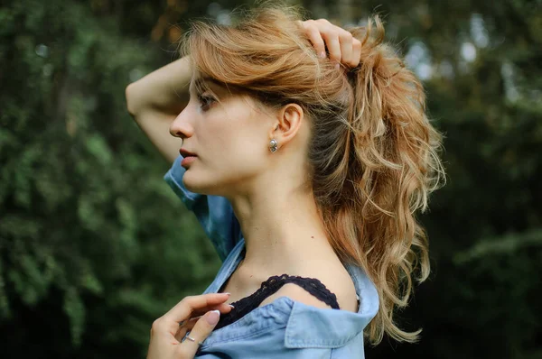 Oldalnézet portré szexi szőke lány göndör haj visel kék alkalmi farmer blúz és fekete fehérnemű pózol a parkban zöld fák háttér. Hipster stílus koncepció. — Stock Fotó