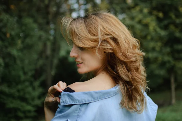 Side view portrait of sexy blonde girl with curly hair wearing blue casual jeans blouse and black lingerie posing in the park on green trees background. Hipster style concept. — Stock Photo, Image