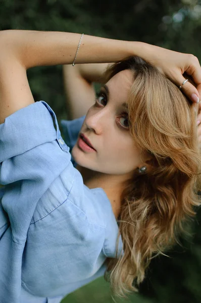 Porträt von sexy blondem Mädchen mit lockigem Haar, das eine blaue lässige Jeansbluse trägt und im Park auf grünem Baumhintergrund posiert. Konzept im Hipster-Stil. — Stockfoto