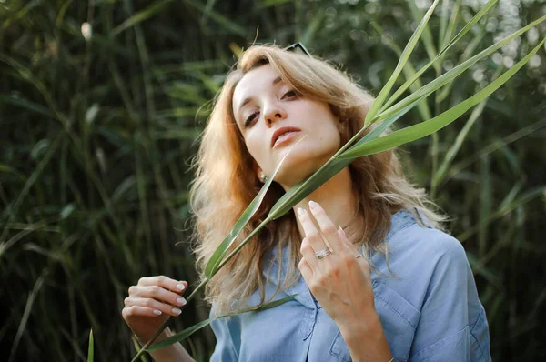 Schöne blonde Mädchen posiert für einen Fotografen in einem blauen lässigen Jeanshemd mit Stiel in der Hand in der Nähe des nackten Halses im Park auf grünem Stielgrund. Konzept im Hipster-Stil. — Stockfoto