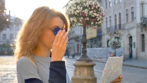 Счастливая взволнованная взрослая девушка смотрит на бумажную карту и ищет направление рано утром в древнем европейском городе на пустой площади — стоковое видео
