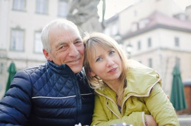 Yakışıklı yaşlı adam, genç sarışın karısını kucaklıyor. Baharın başında ya da sonbaharda antik şehirde birlikte vakit geçiriyorlar..