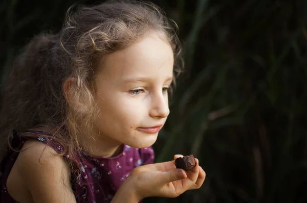 Hermosa niña rubia en vestido violeta con cola de caballo está comiendo un caramelo de chocolate en el parque durante un día, concepto de adicción al azúcar —  Fotos de Stock