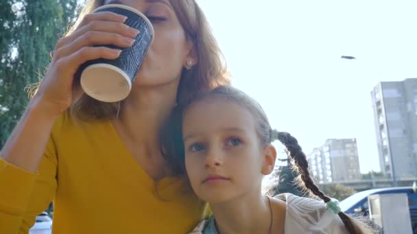 Close-up portret van mooie jonge moeder in geel shirt ontspannen samen met haar dochtertje in de zomerochtend in de buitenlucht cafe, vrouw drinkt koffie uit zwarte papieren beker. — Stockvideo