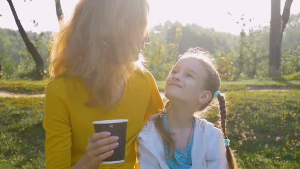 Giovane madre in camicia gialla con tazza di caffè usa e getta e piccola figlia carina si godono la mattina presto seduta alla panchina nel parco sullo sfondo dell'alba — Video Stock
