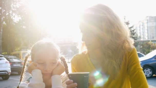 Madre joven en camisa amarilla con taza de café desechable y pequeña hija linda están hablando sentado temprano en la mañana en la terraza de la cafetería en el fondo del amanecer — Vídeos de Stock