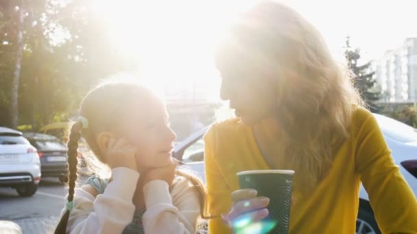 Jonge moeder in geel shirt met wegwerp kopje koffie en kleine schattige dochter praten zitten vroeg in de ochtend op het cafe terras op zonsopgang achtergrond — Stockvideo