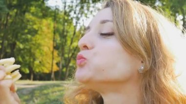 Açık havada, parkta bankta oturmuş patates kızartması yiyen genç sarışın kadının portresini kapat. Sağlıksız yiyecekler, öğle yemeği konseptleri.