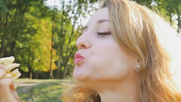 Primer plano retrato al aire libre de una joven rubia comiendo papas fritas sentada en el banco del parque. Alimentación poco saludable, conceptos de la hora del almuerzo — Vídeos de Stock