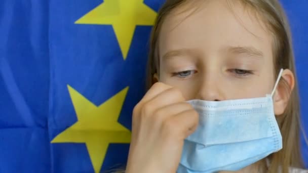 Retrato close-up de menina loira com grandes olhos azuis em máscara disposible para proteção de vírus no fundo bandeira da União Europeia em estúdio. Equipamento de protecção individual — Vídeo de Stock