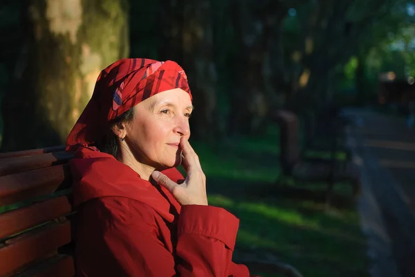 붉은 옷을 입고 공원에 있는 연로 한 피곤 한 여자가 벤치에 앉아 봄이나 가을에 공원에서 좋은 날씨와 일몰을 즐긴다 — 스톡 사진