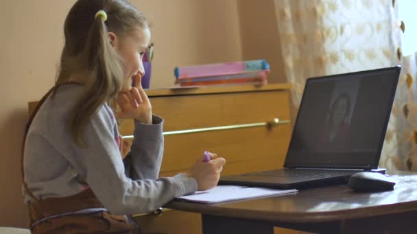 Kleines blondes Mädchen mit zwei Pferdeschwänzen lernt mit ihrem Lehrer per Videochat und schreibt etwas in Schulheft zu Hause wegen der Selbstisolierung durch Coronavirus Covid-19 — Stockvideo