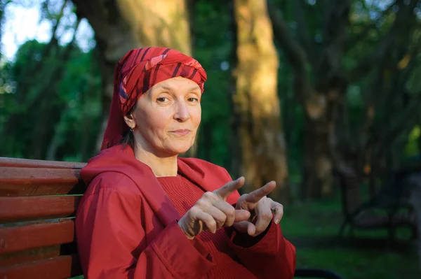 Жіночий портрет зрілої жінки, одягненої в червоний одяг і показує два пальці, що сидять на лавці і насолоджуються хорошою погодою і заходом сонця в парку навесні або восени . — стокове фото