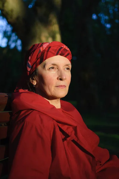 Пожилая уставшая женщина в красной одежде сидит на скамейке и наслаждается хорошей погодой и закатом в парке весной или осенью — стоковое фото