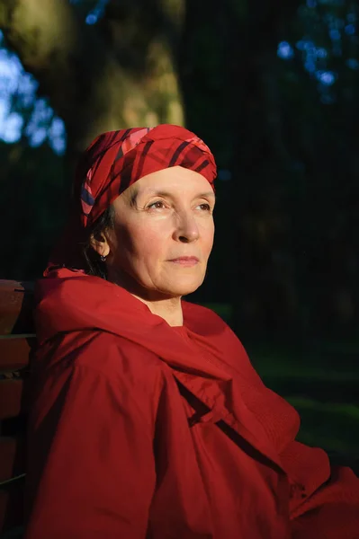 Anciana cansada vestida de rojo en el parque sentada en el banco y disfrutando de un buen tiempo y puesta de sol en el parque durante una primavera u otoño —  Fotos de Stock