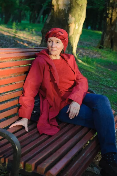 Female portrait of mature woman dressed in red clothes and sitting on the bench and enjoying a good weather and sunset in the park during a spring or autumn. — Stock Photo, Image