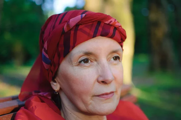 Close up portrait of mature woman dressed in red clothes and sitting on the bench and enjoying a good weather and sunset in the park during a spring or autumn — Stock Photo, Image