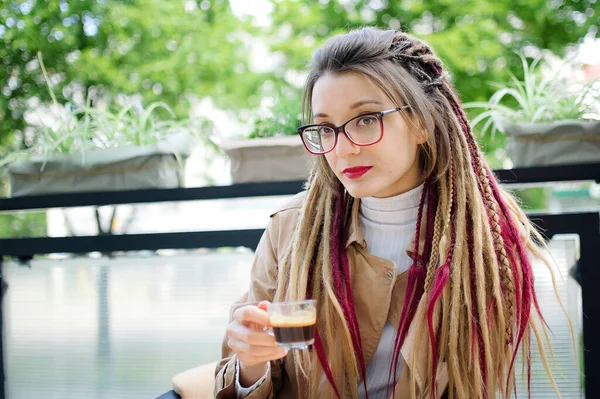 Positive smart student girl with long dreadlocks is resting in a street coffee shop with transparent cup of espresso. — Stock Photo, Image