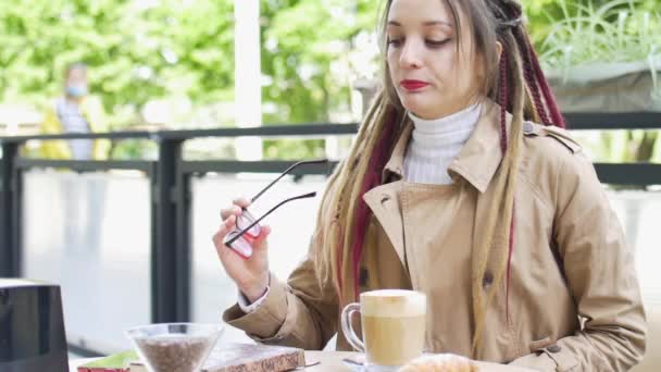 Jeune femme d'affaires moderne avec de longs dreadlocks et lunettes écrit quelques notes dans son carnet tout en étant assis dans un café de rue pendant une journée, concept de travail à distance — Video