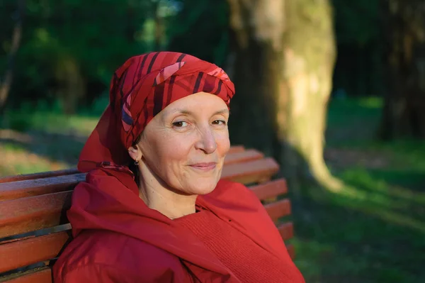 Vrouwelijke portret van volwassen vrouw gekleed in rode kleren en zitten op de bank en genieten van een goed weer en zonsondergang in het park tijdens een lente of herfst. — Stockfoto