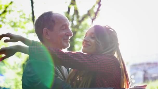 Menina moderna com dreadlocks está abraçando seu pai sênior passar o tempo juntos no parque durante o dia ensolarado. Conceito de dia dos pais — Vídeo de Stock
