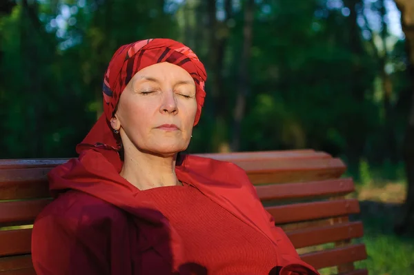 Anciana cansada vestida de rojo en el parque sentada en el banco y disfrutando de un buen tiempo y puesta de sol en el parque durante una primavera u otoño — Foto de Stock